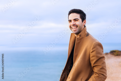 The guy holds his hands in the pockets of a beige coat and laugh