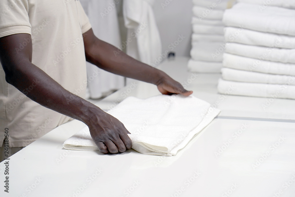 Obraz premium Hands of an african male laundry hotel worker folds a clean white towel. Hotel staff workers. Hotel linen cleaning services.