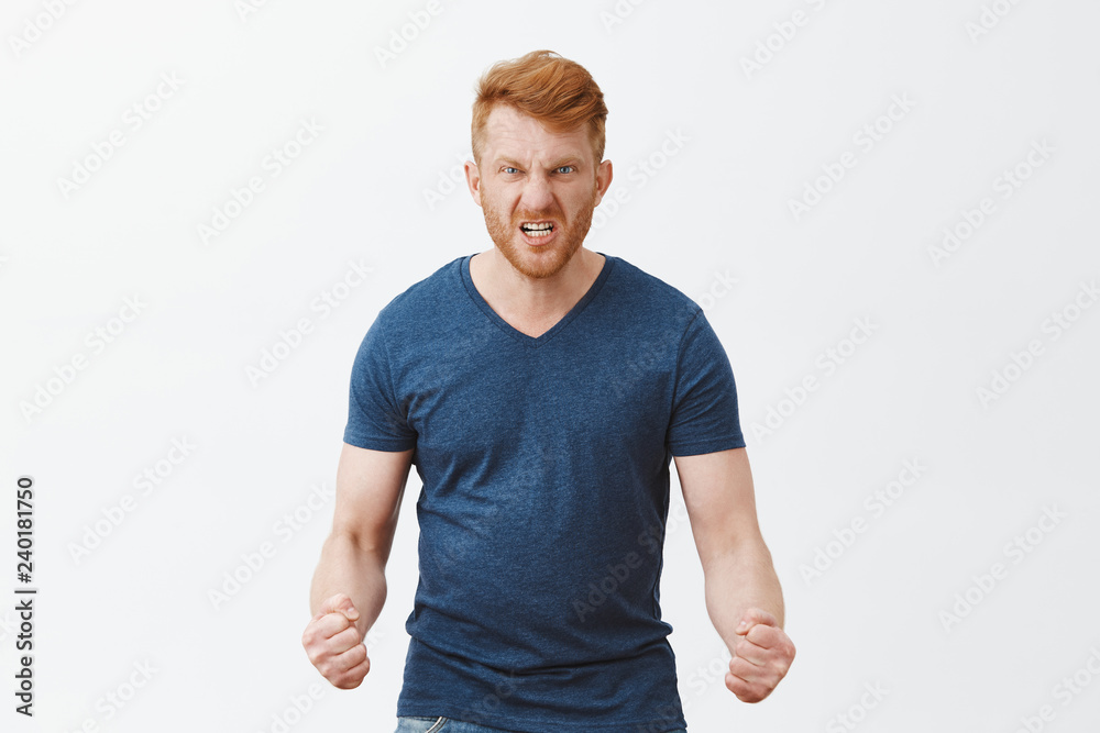 Fototapeta premium Guy will show how anger looks. Portrait of pissed and angry dangerous redhead guy with outraged expression, wrinkling nose, frowning and clenching fists dangerously, feeling hate over gray wall