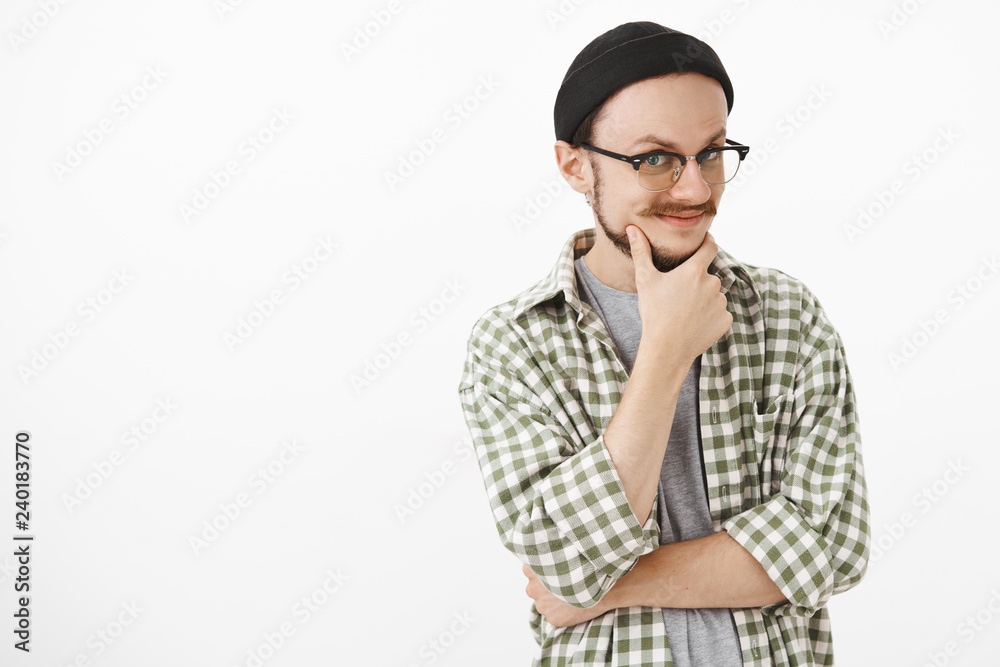 Studio shot of intrigued enthusiastic young creative guy in glasses and black beanie rubbing beard smiling from curiousity and joy gazing mysteriously at camera as if having some idea or intention Studio shot of intrigued enthusiastic young creative guy in glasses and black beanie rubbing beard smiling from curiousity and joy gazing mysteriously at camera as if having some idea or intention