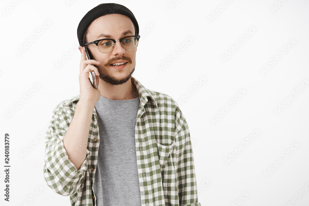 Waist-up shot of carefree calm and relaxed normal guy in beanie and glasses looking right with careless expression while calling friend talking on smartphone holding device near ear Waist-up shot of carefree calm and relaxed normal guy in beanie and glasses looking right with careless expression while calling friend talking on smartphone holding device near ear
