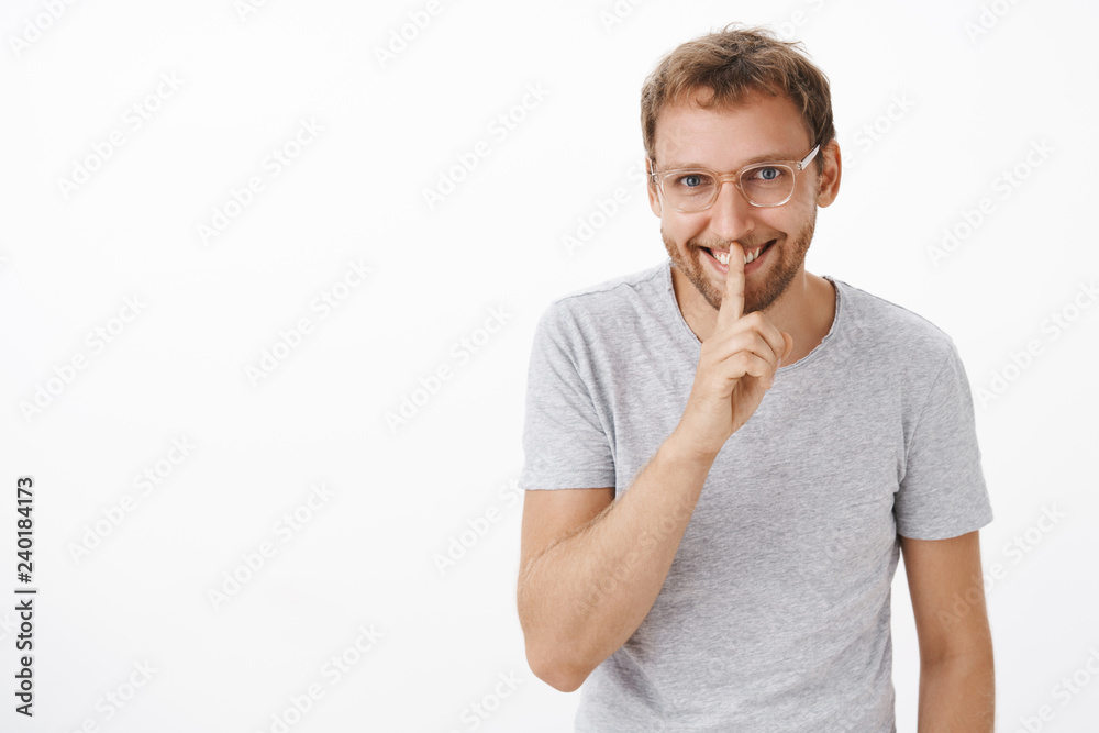 Portrait of mysterious happy and energized handsome man with bristle in glasses and gray t-shirt smiling with enthusiasm saying shh, showing shush gesture with index finger over mouth Portrait of mysterious happy and energized handsome man with bristle in glasses and gray t-shirt smiling with enthusiasm saying shh, showing shush gesture with index finger over mouth