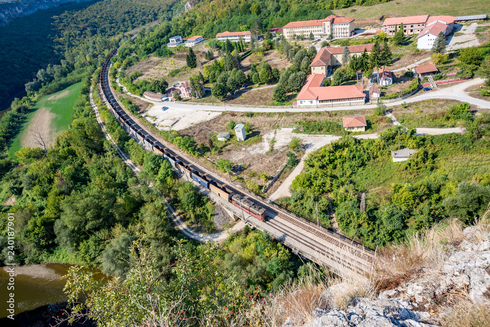 Long empty freight train with many goods wagons passes near the psychiatric hospital near Karlukovo, Northern Bulgaria in sunny autumn day, landscape from high above