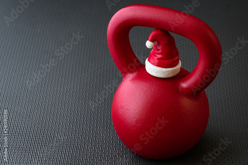 Fototapeta Naklejka Na Ścianę i Meble -  Red kettlebell on a black gym floor with small red and white Santa Claus hat to celebrate Christmas fitness