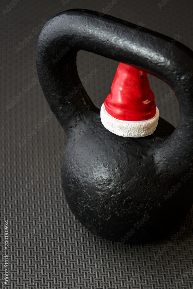 Fototapeta premium Black kettlebell on a black gym floor with small red and white Santa Claus hat to celebrate Christmas