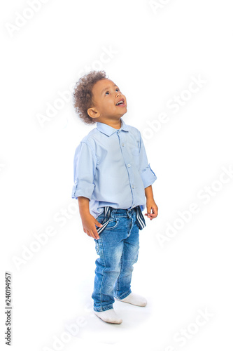 A little dark-skinned boy with curly hair is standing half-turn looking up and smiling pretty, isolated background