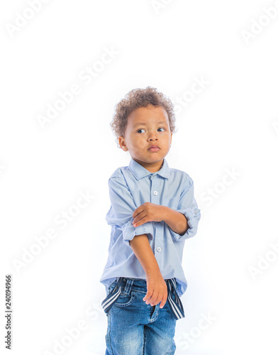 The little dark-skinned boy with curly hair stands holding his sleeve shirt with his hand and looks suspiciously to the side, isolated background