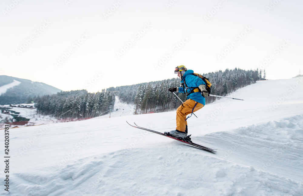 Skier man at jump from the slope of mountains in blue jacket with forest in background. Side view
