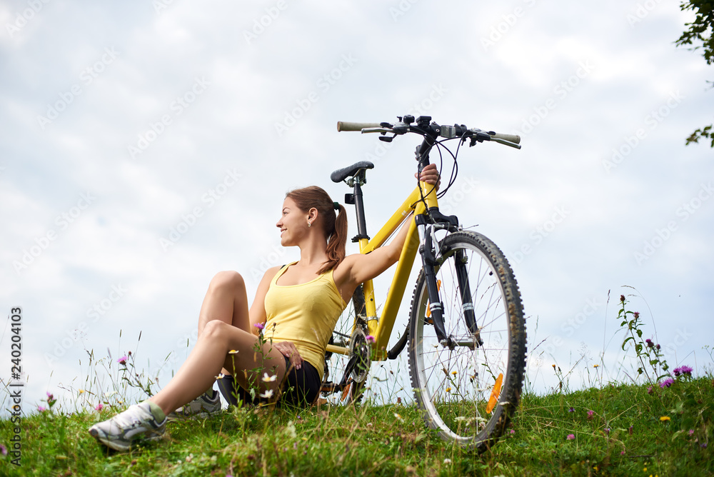 Young happy woman biker sitting on a grassy hill with yellow mountain bicycle, enjoying summer day in the mountains. Outdoor sport activity, lifestyle concept Young happy woman biker sitting on a grassy hill with yellow mountain bicycle, enjoying summer day in the mountains. Outdoor sport activity, lifestyle concept