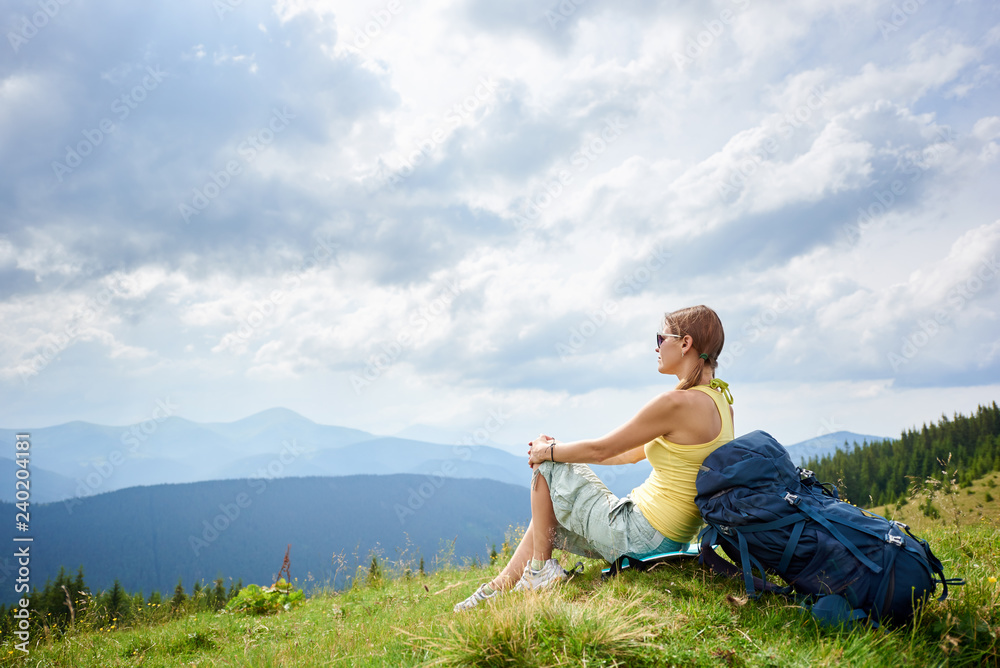 Side view of attractive woman hiker resting on grassy hill with backpack, wearing sunglasses. Female tourist enjoying summer cloudy day in the Carpathian mountains. Outdoor activity, tourism concept
