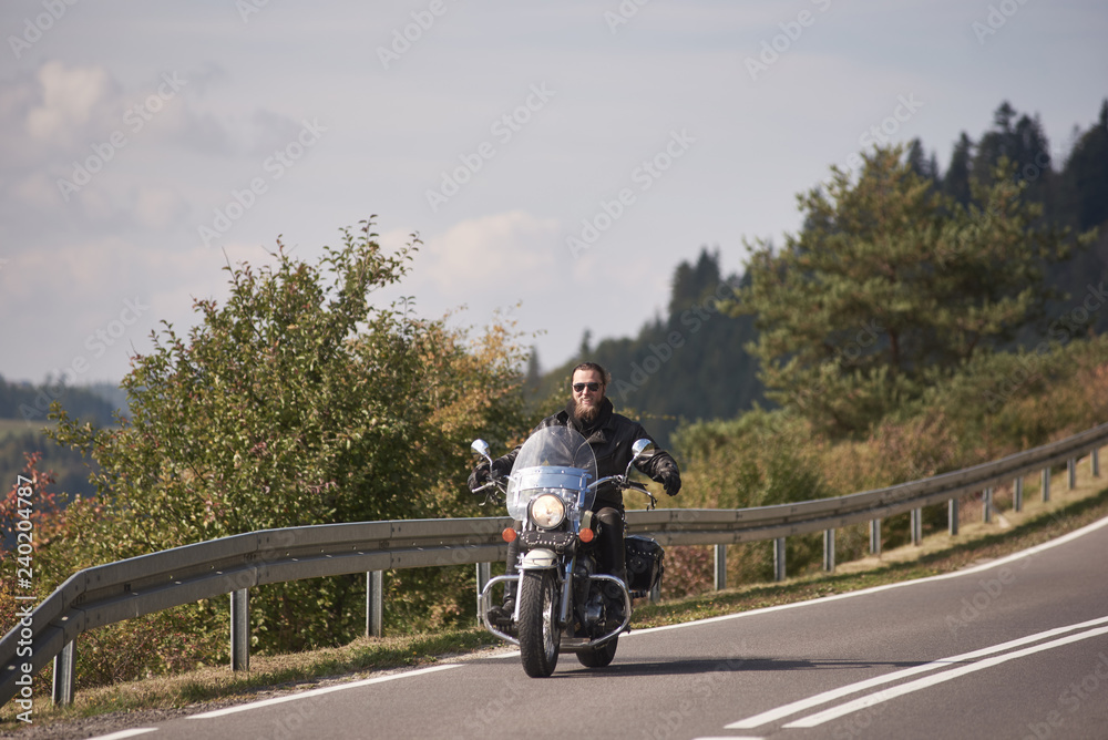 Smiling motorcyclist in black leather outfit and sunglasses riding cruiser motorbike with stretched out arms on bright sunny summer day