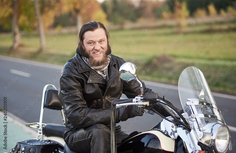Portrait of bearded happy smiling motorcyclist in black leather clothing sitting on cruiser motorbike on blurred vintage bokeh background of golden trees and green grass in the evening.