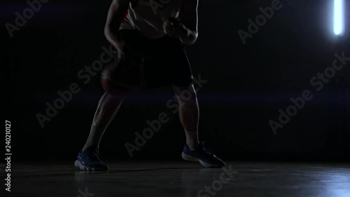 A man with a basketball on a dark basketball court against the backdrop of a basketball ring in the smoke shows dribbling skills illuminated by three lanterns in backlight