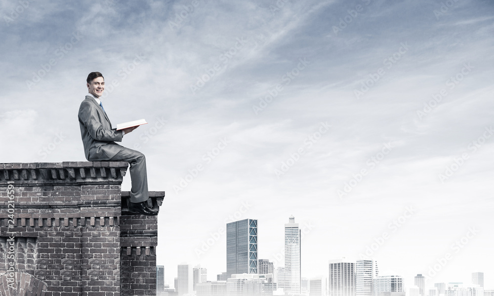 Man on roof edge reading book and cityscape at background Man on roof edge reading book and cityscape at background