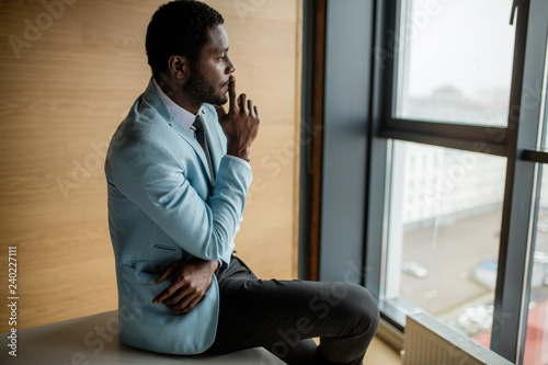 African american businessman in a light blue formal jacket and black pants contemplating cityscape with thoughtful look sitting against big window.