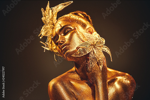 Fashion gold model woman with bright golden sparkles on skin posing, fantasy flower. Portrait of beautiful girl with glowing makeup. Glitter glowing skin, jewellery