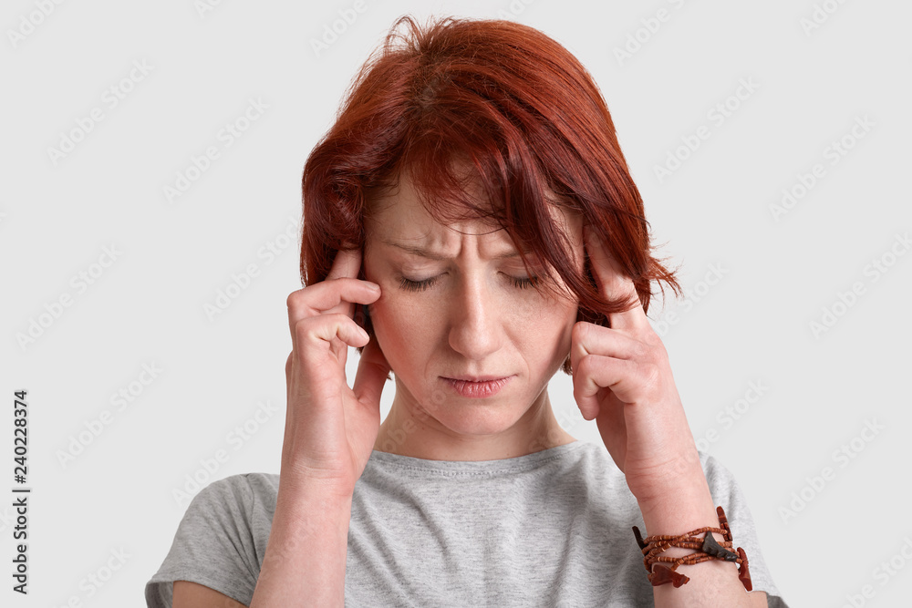 Headshot of stressful red haired young woman keeps both index fingers on temples, suffers from migraine, dressed casually, isolated over white background, tries to find solution. Headahe concept Headshot of stressful red haired young woman keeps both index fingers on temples, suffers from migraine, dressed casually, isolated over white background, tries to find solution. Headahe concept