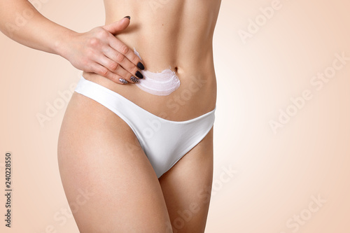 Young fit woman applies cosmetic cream for cellulite or lotion on belly, wears white panties, cares of skin, isolated over beige background, avoids stretching. Cosmetology and medicine concept © sementsova321