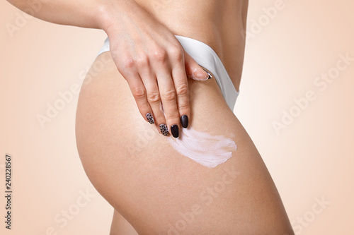 Sideways shot of slim woman applies body cream on hip, wants to have flawless ideal skin, has black manicure, wears white underwear, isolated over beige background. Lady uses lotion for legs.