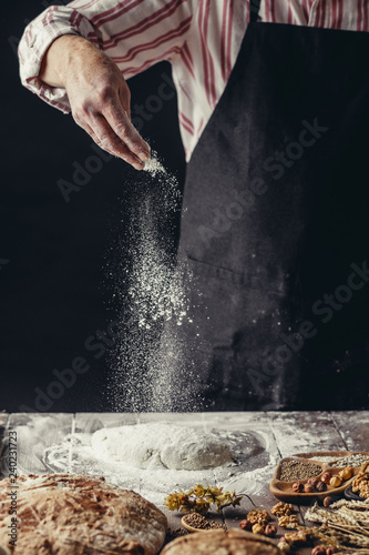 Male Cook hands kneading dough  sprinkling piece of dough with white wheat flour. Low key shot  close up on hands  tasty rustic organic loaf of bread and some ingredients around on table.