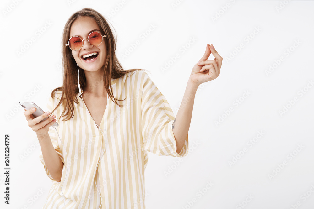 One click and go. Portrait of joyful delighted happy woman in trendy sunglasses and blouse holding smartphone smiling broadly enjoying great music quality in earphones over gray background One click and go. Portrait of joyful delighted happy woman in trendy sunglasses and blouse holding smartphone smiling broadly enjoying great music quality in earphones over gray background
