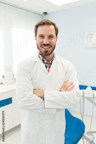 Happy young attractive dentist or doctor smiling looking at camera with arms crossed in his modern dental clinic wearing white uniform. Dentistry and medic concept.