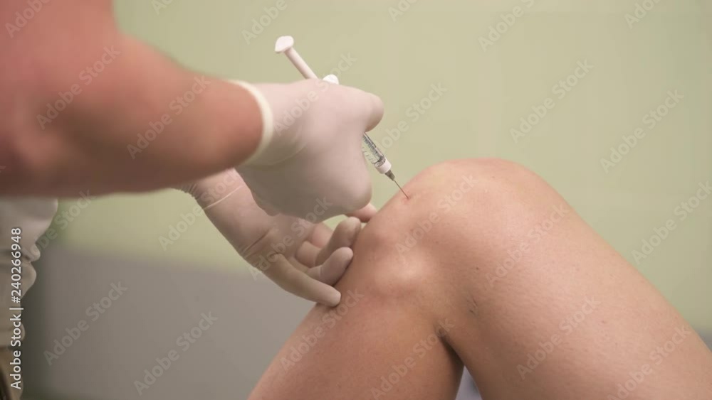 A close-up of the knee, hands in medicine gloves with a syringe start making an injection. Intra-articular injection of hyaluronic acid.