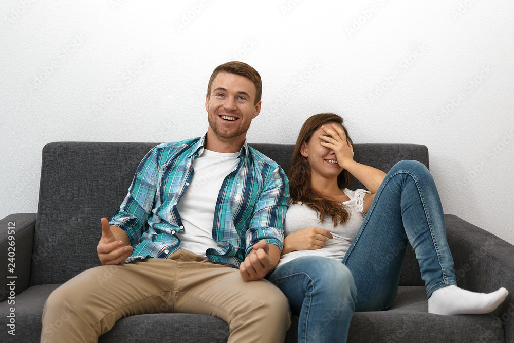 Cute cheerful girl wearing blue jeans covering face and smiling broadly  sitting on sofa with her unshaven husband watching TV. Beautiful European  couple enjoying nice movie on couch at home foto de