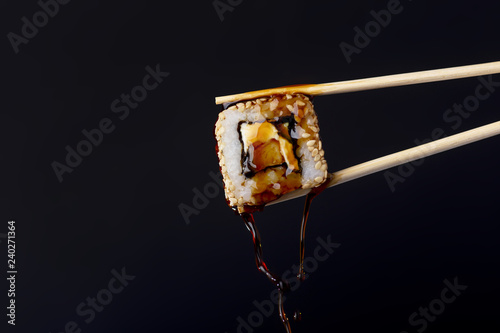 succulent roll between chopsticks on black background, drops of soy sauce dripping from sushi, food background, Japanese cuisine