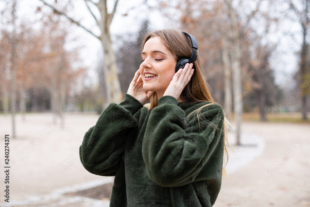 Attractive young female in headphones cheerfully smiling and looking away while walking in autumn park and listening to music Attractive young female in headphones cheerfully smiling and looking away while walking in autumn park and listening to music