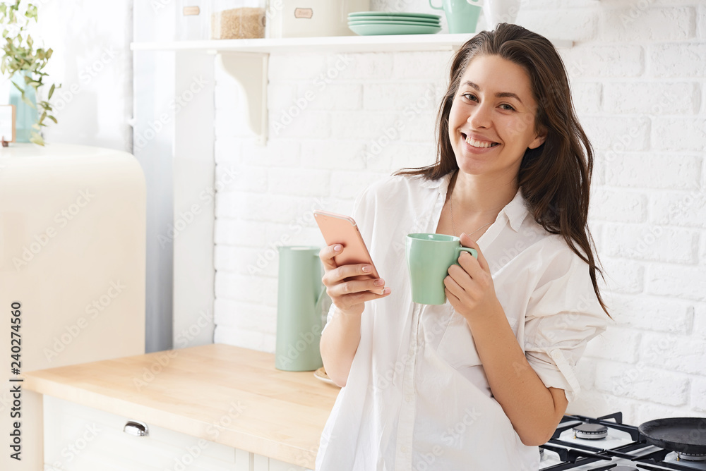 Young woman using smartphone leaning at kitchen table with coffee mug and organizer in a modern home. Smiling woman reading phone message. Brunette happy girl typing a text message.
Young woman using smartphone leaning at kitchen table with coffee mug and organizer in a modern home. Smiling woman reading phone message. Brunette happy girl typing a text message.
