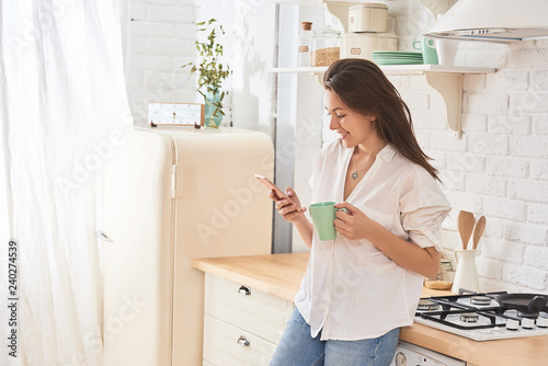 Young woman using smartphone leaning at kitchen table with coffee mug and organizer in a modern home. Smiling woman reading phone message. Brunette happy girl typing a text message.

