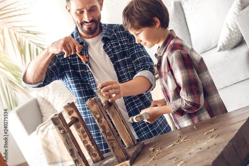 Father and little son at home standing dad screwing screw into chair cheerful while boy holding hammer