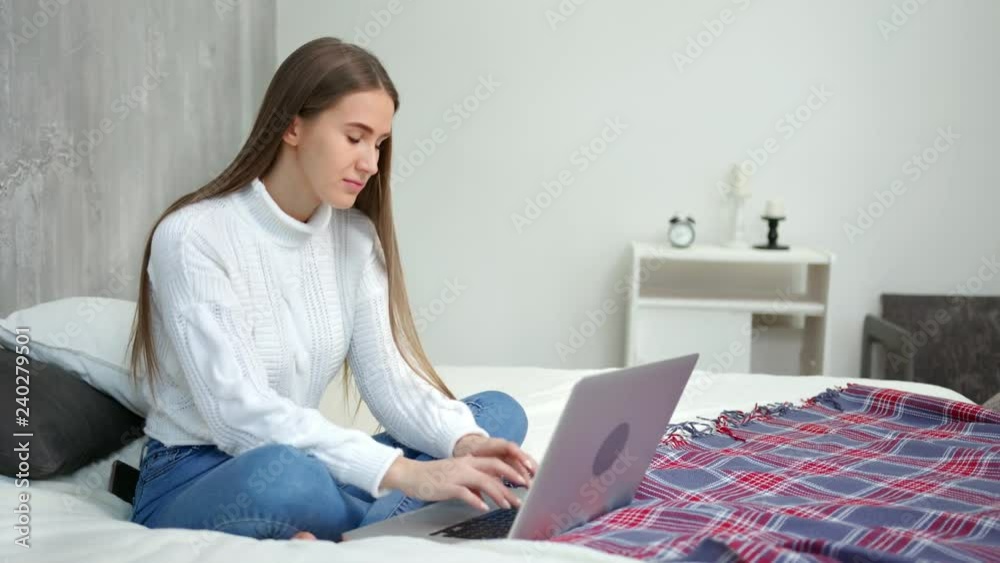 Focused smiling young freelancer woman working using laptop sitting on bed at cozy home Focused smiling young freelancer woman working using laptop sitting on bed at cozy home