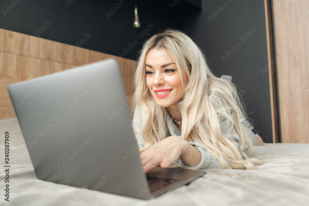 portrait of a girl behind a laptop screen. Beautiful blonde woman lying on the bed and looks at the laptop screen. Blogging, browsing internet, chatting. In a warm cozy sweater portrait of a girl behind a laptop screen. Beautiful blonde woman lying on the bed and looks at the laptop screen. Blogging, browsing internet, chatting. In a warm cozy sweater