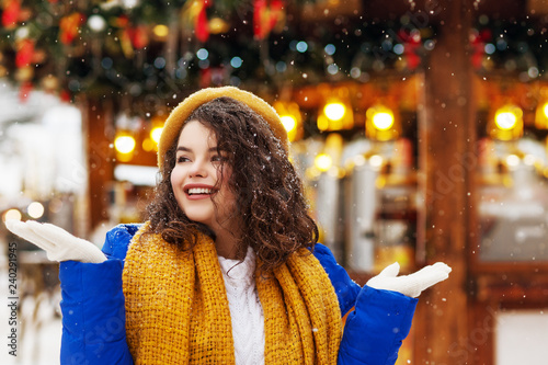 Outdoor portrait of young happy smiling lady catching snowflakes, posing at festive Christmas fair in european city. Model wearing yellow beret, scarf, white knitted sweater, gloves, blue winter coat photo