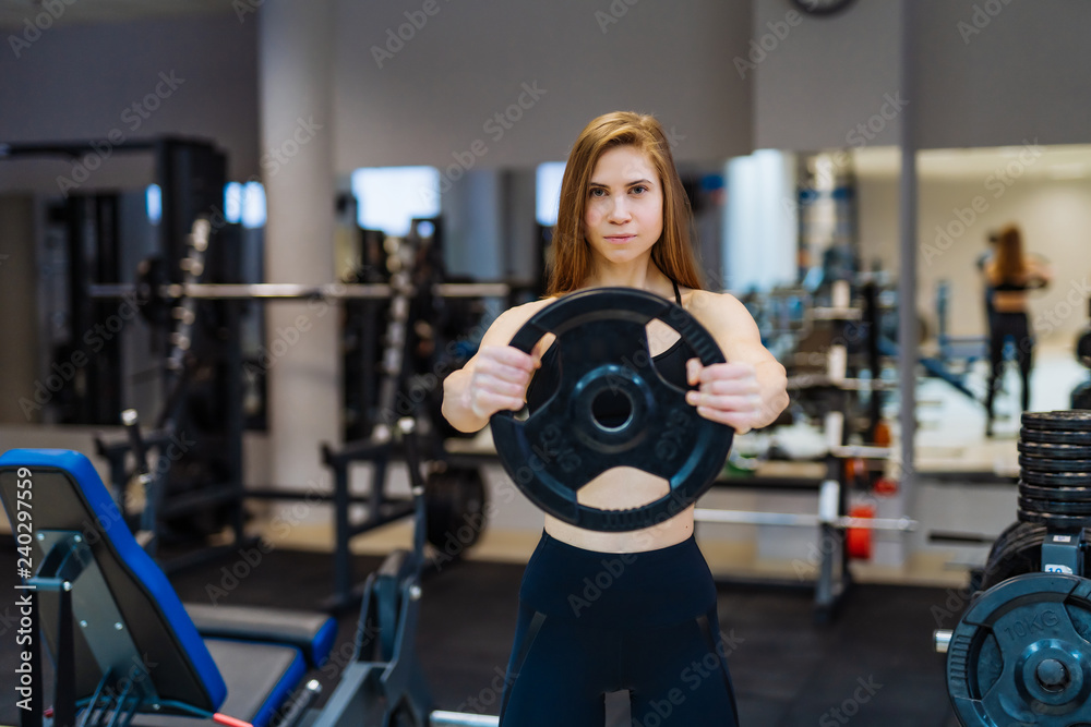 Seductive fitness woman in sportswear posing with a barbell disk at gym. Professional sport fitness female with weight disk in gym.