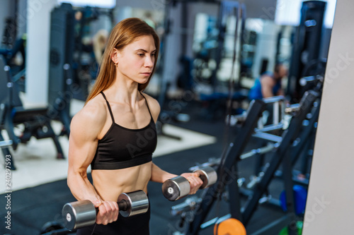 Strong athletic woman in black sport wear pumping up muscles with dumbbells in the gym. Fitness girl with perfect body in the gym has a training.