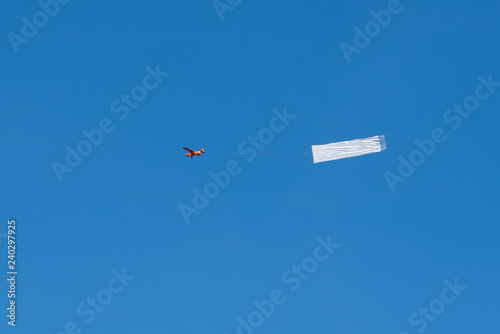A small red airplane is pulling a white banner in the blue sky.
