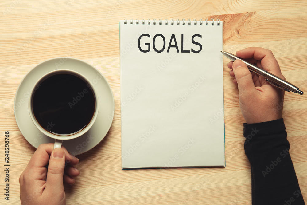 Text GOALS on notepad on wooden table and female hands with pen and coffee, top view Text GOALS on notepad on wooden table and female hands with pen and coffee, top view
