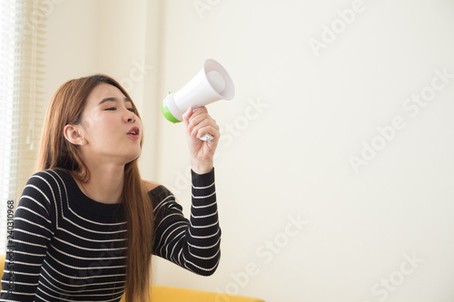 Beautiful young Asian woman making announcement or communicates with megaphone, Message announce from teenager wearing a black shirt and sitting on yellow sofa in the living room at home