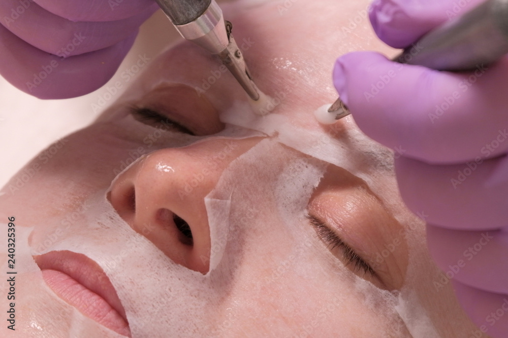 Fototapeta premium Close-up of electrodes in the hands of a beautician, touching the female cheek through a mask. Woman receiving stimulating facial treatment with microcurrent equipment. Wrinkle smoothing.