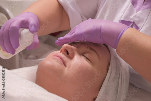 A professional cosmetologist in lilac gloves with one hand covers his eyes, while the other sprays a moisturizing tonic on the face of a middle-aged woman.
