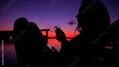 Silhouette of Young Unidentified Woman Smoking Weed Joint or Cigatette Againts Amazing Beach Sunset near Water. 4K Slowmotion. photo