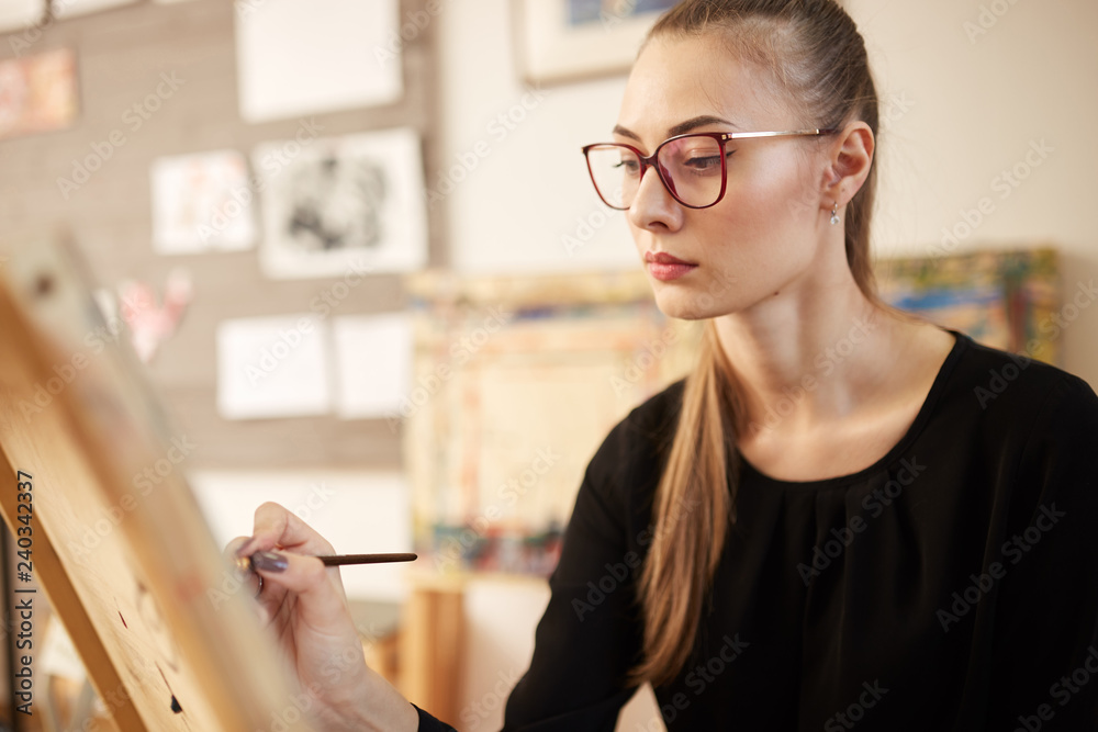 Charming fair-haired girl in glasses dressed in black blouse sits at the easel and paints a picture in the art studio Charming fair-haired girl in glasses dressed in black blouse sits at the easel and paints a picture in the art studio