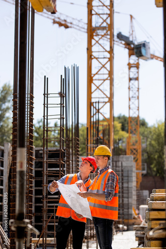 Construction manager and engineer dressed in orange work vests and hard helmets explore construction documentation on the building site near the steel frames