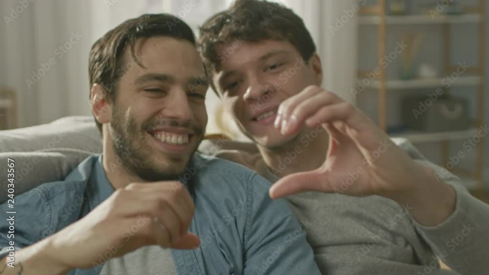 Portrait of a Cute Male Queer Couple at Home. They Sit on a Sofa and Look at Each Other. Partner Has One Hand Behind His Lover. They Make a Heart Symbol with Their Hands. Room Has Modern Interior.
