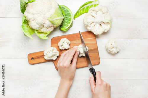 Female hands cut cauliflower with knife on cutting board, kitchen towel on rustic white wooden background top view flat lay copy space. Cooking, healthy wholesome food, vegetable, diet concept.