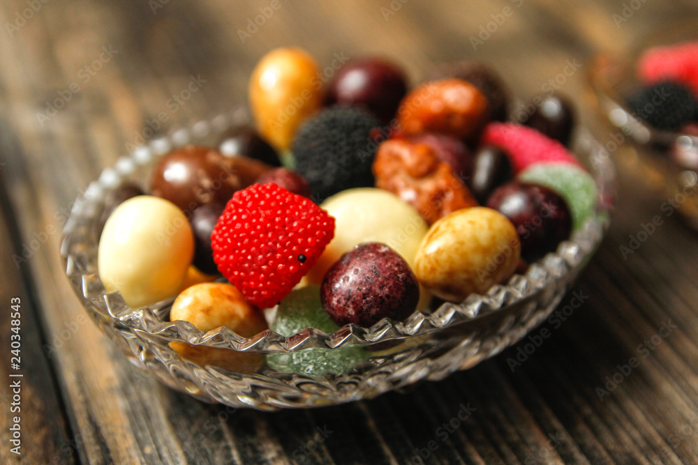 Different candies in a crystal bowl on old wooden table. Multi colored gummy candy coated with sugar, chocolate and fruit candy. Different candies in a crystal bowl on old wooden table. Multi colored gummy candy coated with sugar, chocolate and fruit candy.