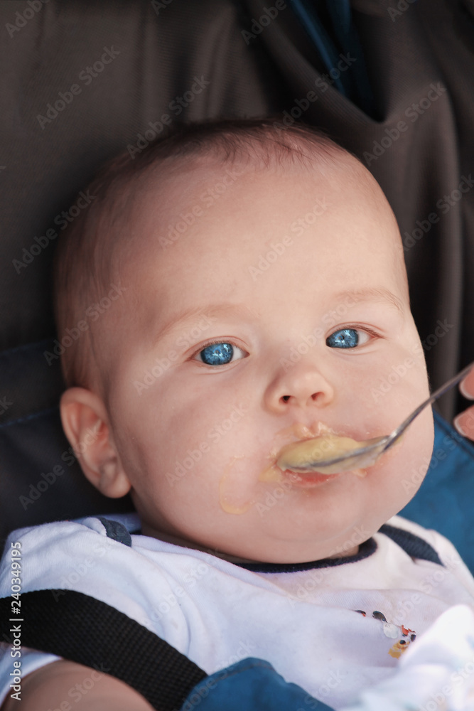 Cute caucasian baby boy fed from small spoon in baby chair. Close up portrait of pretty baby with bright blue eyes. Outdoors, copy space. Cute caucasian baby boy fed from small spoon in baby chair. Close up portrait of pretty baby with bright blue eyes. Outdoors, copy space.